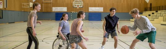 Fünf Jugendliche spielen in einer Sporthalle Basketball.