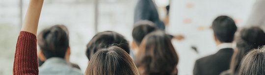 Menschen sitzen in einem Seminar. Eine Frau meldet sich. Es steht "Noch Plätze frei" im Bild. 